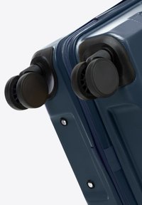 Valigia rigida di colore blu navy con superficie texturizzata, dotata di due ruote nere e una cerniera, con un manico incassato sul lato.