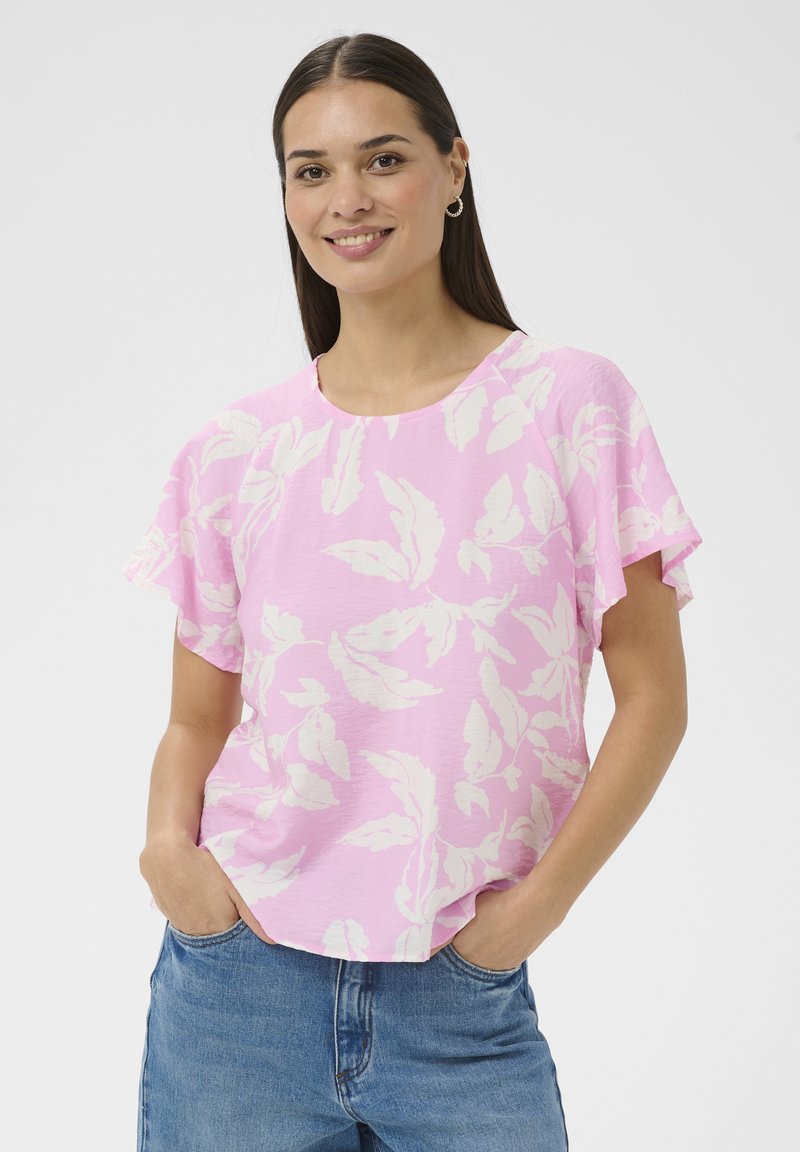 Femme souriante aux longs cheveux foncés portant des boucles d'oreilles créoles, un chemisier rose à fleurs à manches courtes et un jean bleu, mains dans les poches, sur fond blanc.