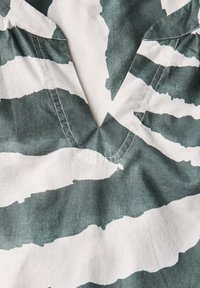 Tissu en coton avec un motif abstrait de vagues vertes et blanches. Présente un col en V et des détails à volants aux épaules. Coutures visibles en accent.