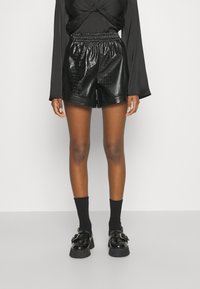 Shorts noirs en faux cuir à texture crocodile, taille élastique, longueur mi-cuisse, associés à des chaussettes noires aux chevilles et des mocassins épais.