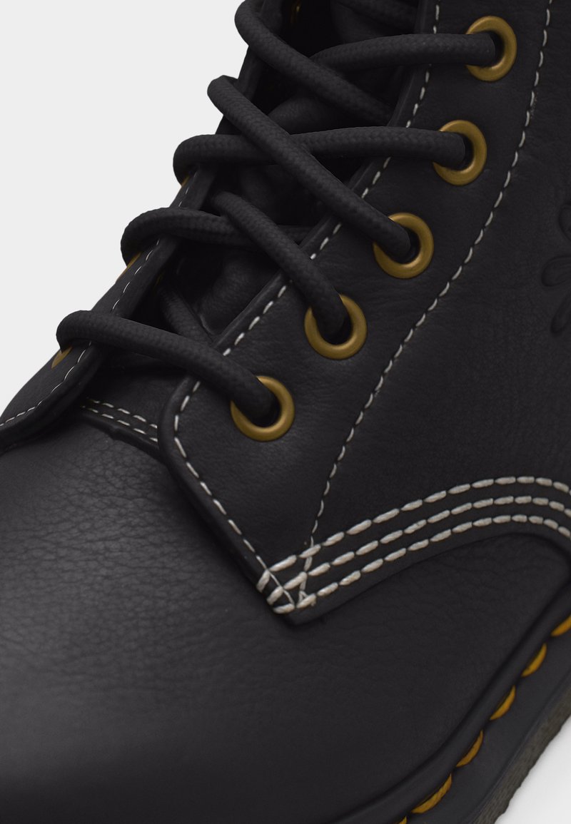 Gros plan sur une botte en cuir noire avec des lacets noirs, des œillets métalliques jaunes et des détails de couture blancs.