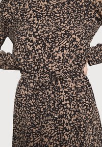 Klänning i svart och beige mönstrad tyg med cinchad midja, justerbar dragsko och långa ärmar med knutdetaljer.