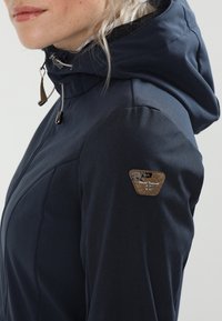 Marineblaue wasserdichte Jacke mit tailliertem Schnitt, hohem Kragen, verstellbarem Kapuzenansatz und einem kleinen braunen Logo-Patch auf der linken Schulter.