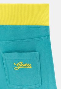 Tessuto turchese con una cintura gialla a coste, caratterizzato da una piccola tasca posteriore ricamata con "Guess" in giallo. Texture liscia e design casual.
