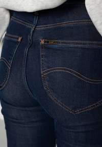 Mörkblå denimjeans med en jämn textur; de har rundade bakfickor och synlig orange sömnad samt en liten "Lee"-logotyp.
