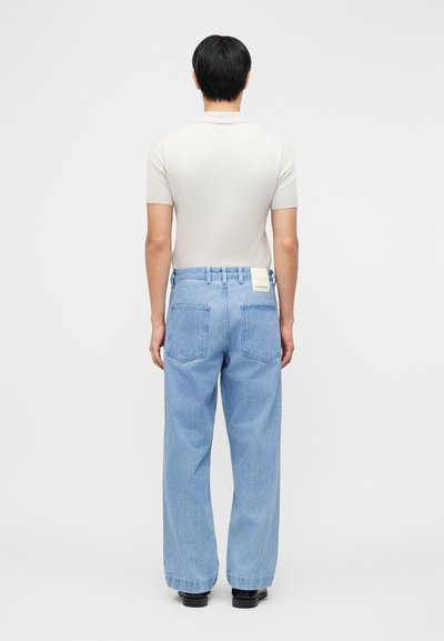 Άνδρας που στέκεται προς τα πίσω φορώντας μπεζ πουκάμισο με κοντά μανίκια, ανοιχτό μπλε τζιν με φαρδιά πόδια και μαύρα παπούτσια, μπροστά σε λευκό φόντο.