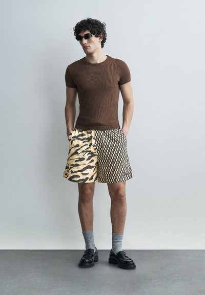 Jeune homme portant un t-shirt marron ajusté, un short à motifs avec des dessins de tigre et de damier, des chaussettes grises, des chaussures noires et des lunettes de soleil foncées, debout à l'intérieur.