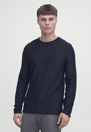 Ung mand med kort hår iført en mørk tekstureret langærmet skjorte og grå bukser, stående mod en ensartet lys baggrund.