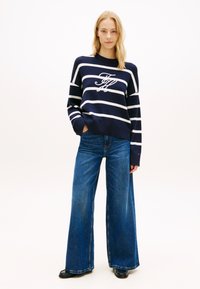 Μπλε πουλόβερ με λευκές ρίγες και λογότυπο, σε συνδυασμό με μπλε τζιν παντελόνι φαρδιάς γραμμής. Λείο ύφασμα και άνετη εφαρμογή.