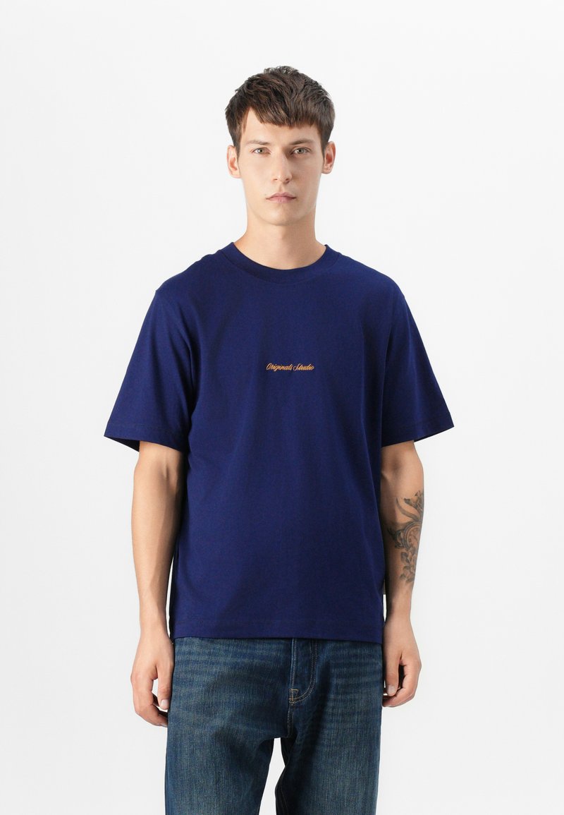 Marineblå bomulds t-shirt med rund halsudskæring og korte ærmer, der har et lille tekstoptryk i guld på brystet. Bæres med et par blå jeans.
