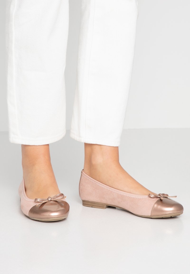 Pieds portant des ballerines rose clair avec embouts métalliques et petits nœuds, associées à un pantalon blanc court sur un sol uni.