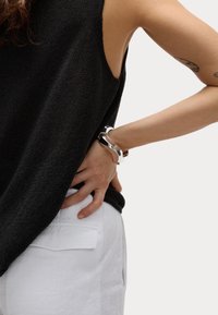 Top sin mangas de punto negro, pantalones de lino blancos, pulsera de plata en la muñeca, mano en la cadera. Detalle del tejido de punto texturizado visible.