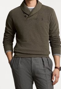 Homme portant un pull col châle vert olive avec des boutons noirs, superposé sur une chemise grise, associé à un pantalon plissé gris et une montre-bracelet marron.