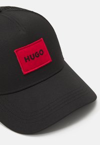 Svart caps laget av stoff med en rød rektangulær lapp med "HUGO" i svart skrift. Den har en buet skjerm og detaljert søm.