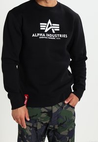 Svart sweatshirt med vitt "Alpha Industries"-logotyp och grafik på framsidan; ribbade muddar och nederkant; bärs med kamouflage cargo-byxor.