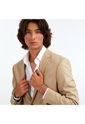 Beige blazer med texturerat mönster, vit skjorta under och silverarmband på handleden. Modellen poserar med händerna på slaget.
