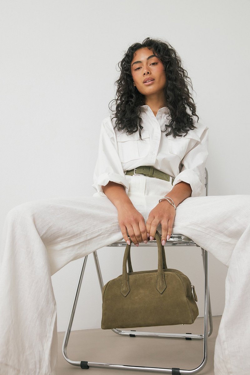 Sac à main en daim vert olive avec deux poignées, forme arrondie, design simple. Modèle assis en chemise blanche et pantalon large sur fond neutre.