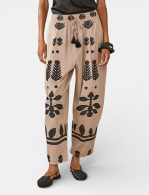 Pantaloni beige larghi con stampe floreali astratte nere, vita elastica con nappine, indossati con top nero, braccialetto nero e sandali neri.