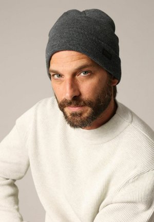 Bonnet gris tricoté avec une texture côtelée et une étiquette, associé à un pull beige crème avec des côtes verticales.