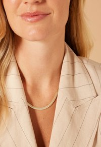 Goudkleurige gelaagde ketting gedragen over een lichtgekleurde, gestreepte blazer. De blazer heeft een schouderkraag en een soepele stofstructuur.