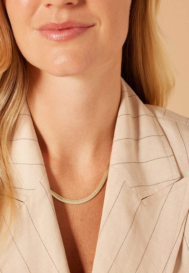 Goudkleurige gelaagde ketting gedragen over een lichtgekleurde, gestreepte blazer. De blazer heeft een schouderkraag en een soepele stofstructuur.