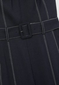 Marineblauwe stof met witte strepen, voorzien van een brede riem met een vierkante gesp en gestikte accenten voor extra textuur en detail.