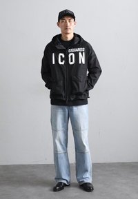 Svart hettegenser med "ICON"-tekst på framsiden, båret over en svart topp. Lys blå slitte jeans og svarte sko er også synlige.
