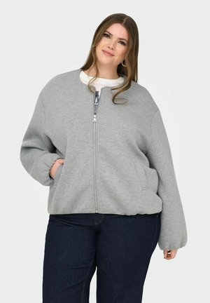 Frau trägt einen grauen Sweatshirt mit Reißverschluss und Taschen sowie dunkle Jeans, steht mit einer Hand in der Tasche vor einem einfarbigen Hintergrund.