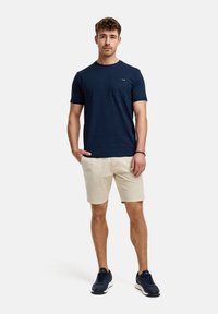 Navy blauwe crewneck t-shirt met een borstzak, gecombineerd met lichtbeige shorts en zwarte sportschoenen. Casual fit en lichtgewicht stof.