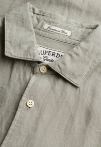Chemise en lin gris clair avec col boutonné, deux boutons visibles et étiquettes en tissu portant les inscriptions "Laundered Wash" et "Superdry".
