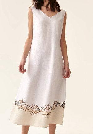 Ærmeløs hvid midi-kjole med V-hals, med et brunt bladmønster langs nederste kant, båret af en stående person.