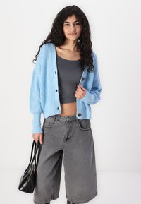 Lichtblauwe gebreide cardigan met een oversized pasvorm, met knoopdetails, gedragen over een donkergrijze cropped top en grijze wijde jeans. Zwarte handtas.