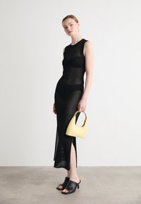 Rochie din plasă neagră cu siluetă ajustată, fante pe lateral, purtată cu tocuri negre cu vârful deschis. Ține în mână o geantă mică galbenă, cu textură netedă.