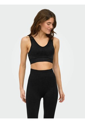 Femme portant un crop top noir sans manches et un legging taille haute, debout contre un fond clair uni, regardant vers le bas et sur le côté.
