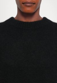 Lähikuva henkilöstä, joka käyttää mustaa pörröistä neuleta, jossa on ribbireunus kaula-avauksessa, näyttäen heidän alaleukaansa ja kaulaansa.