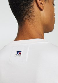 Vit bomullströja med ribbad halsringning och en fyrkantig logotypapplikation med blå, röd och vit accent på baksidan.