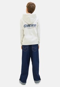 Sweat-shirt gris avec capuche, affichant un texte bleu dans le dos ; associé à un pantalon en denim bleu large, retroussé aux poignets.