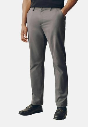 Mann trägt graue maßgeschneiderte Hose, schwarze Loafers und ein schwarzes Hemd, steht mit einer Hand in der Tasche und der anderen entspannt an der Seite.