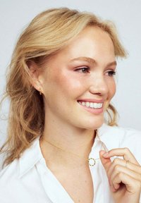 Guldhalsband med en snurrad cirkelpendant, bärs på ett fint kedja. Modellen har blont hår och bär en vit skjorta med knappar.