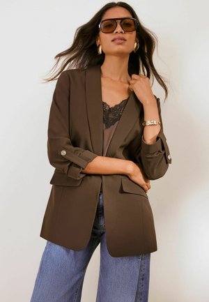 Frau trägt übergroßen braunen Blazer, schwarzen Spitzen-Top, blaue Jeans, große Sonnenbrille, goldene Creolen und ein goldenes Armband vor einfarbigem Hintergrund.