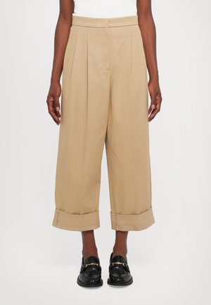 Beige, high-waisted trousers with pleats, wide legs, and rolled cuffs. Smooth fabric, paired with black loafers featuring gold accents.