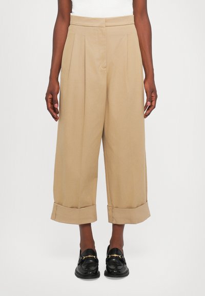 Pantaloni beige a vita alta con pieghe, gamba larga e risvolti. Tessuto liscio, abbinati a mocassini neri con dettagli dorati.
