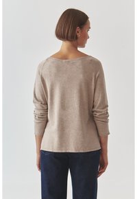 Beige Strickpullover mit langen Ärmeln, weitem Ausschnitt und hochgekrempelten Ärmeln, aus einem strukturierten Strickstoff gefertigt. Getragen mit dunkelblauen Jeans.