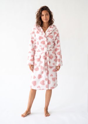 Femme debout pieds nus portant un peignoir blanc à capuche avec des motifs de cœurs roses noué à la taille sur un fond blanc uni.