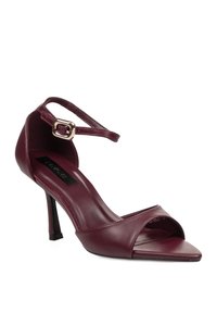 Sandalia de tacón alto de cuero color burdeos con punta afilada, correa en el tobillo y hebilla dorada sobre fondo blanco.