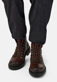 Persona con jeans blu scuro e stivali da trekking in pelle marrone con suole nere, mostrata dalla metà del polpaccio in giù su sfondo bianco.