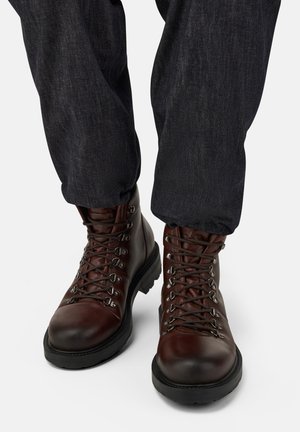 Person i mørkeblå jeans og brune lædervandrestøvler med sorte såler, vist fra midt på læggen og ned mod en hvid baggrund.