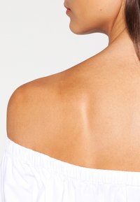 Top bianco off-the-shoulder con un colletto elasticizzato arricciato. Colore della pelle liscia visibile sulla spalla e parte della schiena.