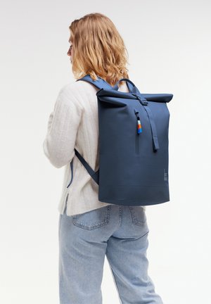 Rolltop-Rucksack in Marineblau, aus strapazierfähigem Material gefertigt. Mit einem vorderen Reißverschluss, verstellbaren Trägern und einem kleinen bunten Akzent.