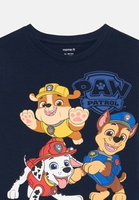 Tri kreslené psy oblečené ako hasič, stavbár a policajt s textom "PAW Patrol" na tmavomodrom tričku.